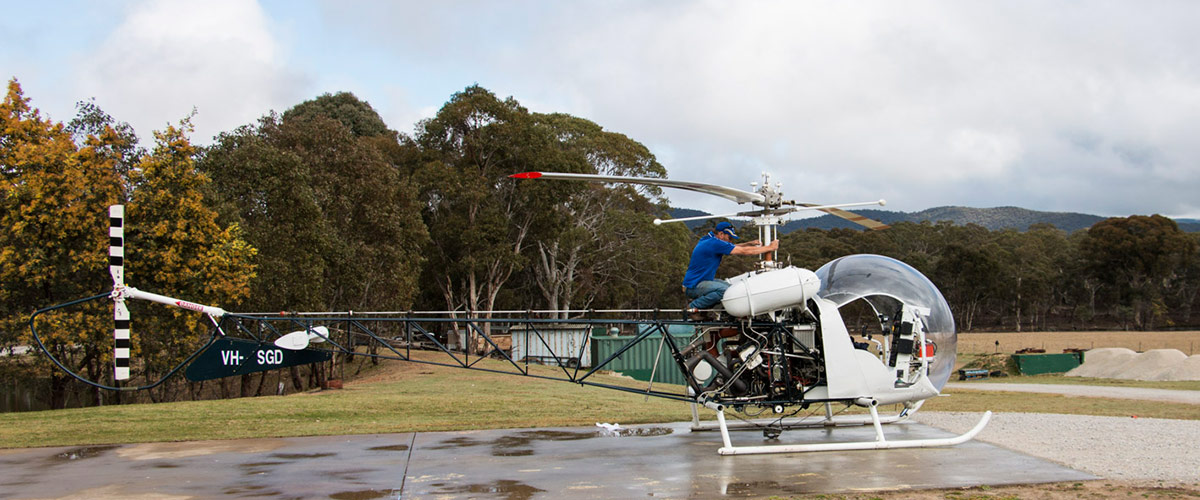 Capertee Valley Helicopter VH-SGD on landing field having pre-flight check of Main rotor by pilot who is sitting atop of the forward part of tail frame. In the background are containers trees and hills.