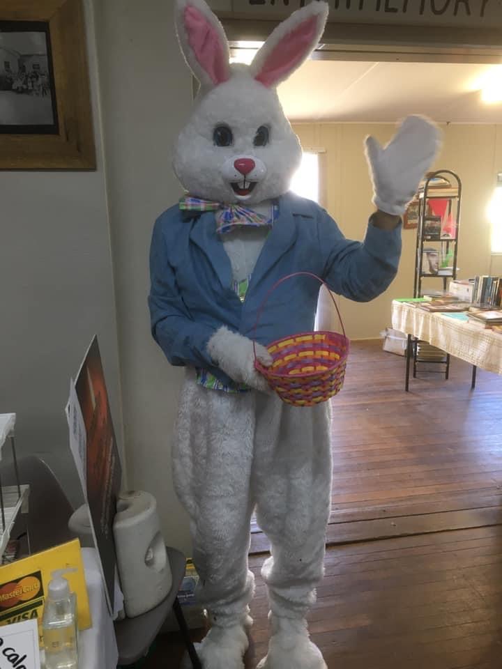 Person Dressed in an Easter bunny suit with blue jacket and waving with one hand and holding a basket in the other. The picture was taken in the Capertee memorial hall.